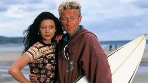 Carl Prechezer/FilmFour A young Catherine Zeta-Jones, who is wearing a flowery dress over the top of a striped T-Shirt, posing with Sean Pertwee, who has a surfboard under his arm, on a beach.