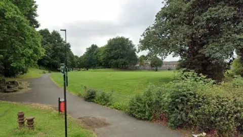 The entrance to a park. A footpath snakes ahead, flanked by open grass and large trees. Lamp-posts also line the route.