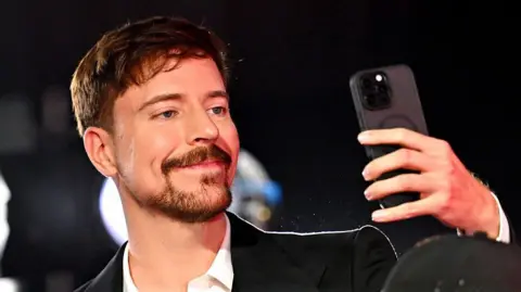 Getty Images MrBeast wearing a black suit and white shirt, smiling while taking a selfie on a black phone.