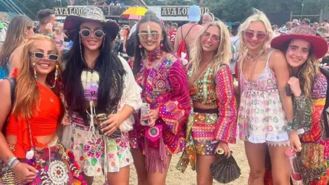 Six young women wearing colourful bohemian-style clothes smile at the camera as they stand in a field at Glastonbury.