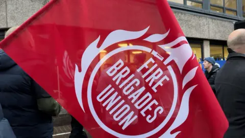 A large red flag displays the emblem of the Fire Brigades Union in white. The name is ringed by a fire hose with a bell above and flames around it. Bahind the flag people are gathered outside the Oxfordshire County Council offices.