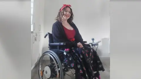 Francesca Lane A woman with dark hair with a red ribbon in it, wearing a red top and a black patterned skirt, sitting in a wheelchair and smiling at the camera.