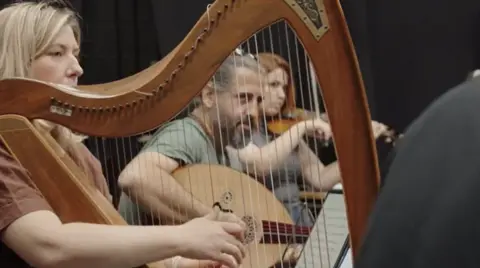 Musicians rehearse together, playing a mix of stringed instruments including a large wooden harp, a lyre‑style instrument, and a violin. Sheet music is visible in the foreground, and the group is seated in a rehearsal space.
