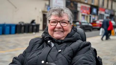 Edna, wearing a black jacket, photographed on Greenock's main shopping high street. She is a pensioner with short grey hair, black glasses and is sitting in an electric mobility scooter. There are small shops and convenience stores behind her, as well as council blue and black bins lined up along a wall.