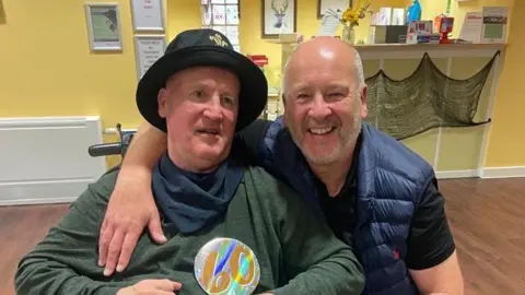 Anthony has grey hair and beard in a blue gilet with a black t-shirt underneath. He has his arm round his brother Keith who is in a wheelchair and is wearing a green long-sleeved t-shirt with a navy bandana round his neck and a black hat on.