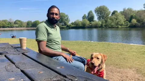 Chandru Balasubramaniam sits at a slightly melted picnic bench with his dog and coffee. he is smiling at the camera on a sunny day with the Coate water lake in the background.