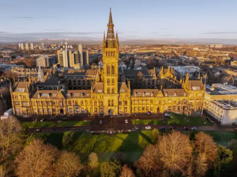 Getty Images Aerial view of the University of Glasgow's main Gilbert Scott Building, with its prominent central tower and Gothic architecture, overlooking the surrounding city skyline in low winter sunlight.