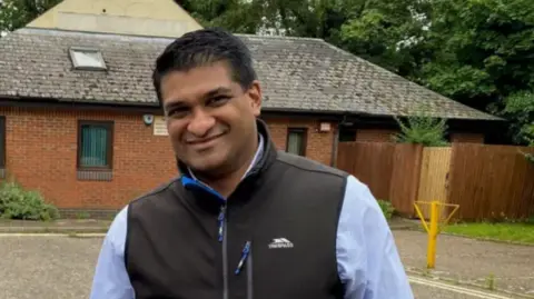 West Suffolk Council Indy Wijenayaka wearing a gillet over a light blue shirt. He is looking into the camera while smiling.