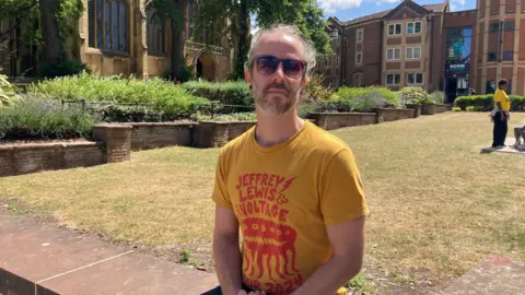 A man with wavy white hair sits on a low wall surrounding a grassy area. He is wearing a yellow T-shirt with some red writing on and sunglasses. There is a church and some buildings surrounding the green behind him.