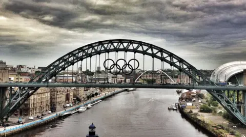 The Tyne Bridge, over the River Tyne, is a curved green metal road bridge. It shows it adorned with the Olympic rings during London 2012. 