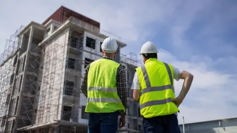 Getty Images Two engineers wearing hardhats and high viz are inspecting the specifications of buildings, apartments, and residences
