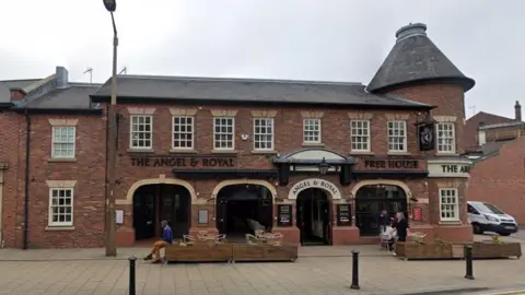 Google Maps A red brick pub with the lettering "The Angel & Royal Free House" above the four entrance arches. Several customers can be seen sitting in chairs on the right.  A man sits on a planter on the left with his legs crossed.