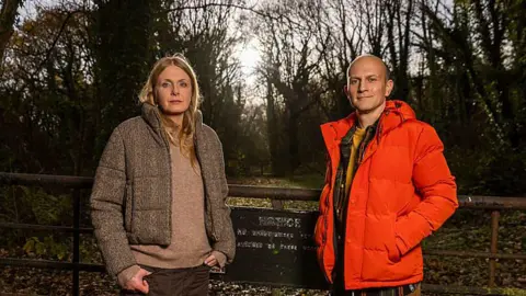 BBC/Wall to Wall Media/Stephen Perry Two people pose beside a countryside gate leading into a wooded path. Winter sunlight filters through leafless trees behind them, while the gate and weathered sign serve as the focal point of the woodland entrance.