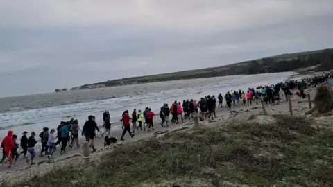 Line of people running on the sand along a beach with grass and sand to the right and the sea to the left.