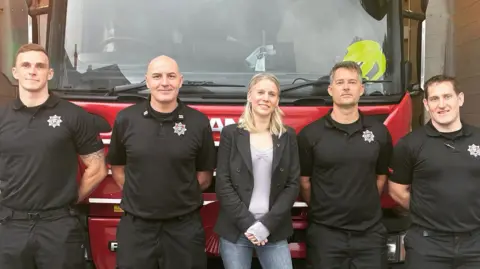 A woman stood with two men on either side of her. They are all stood in front of a fire engine.
