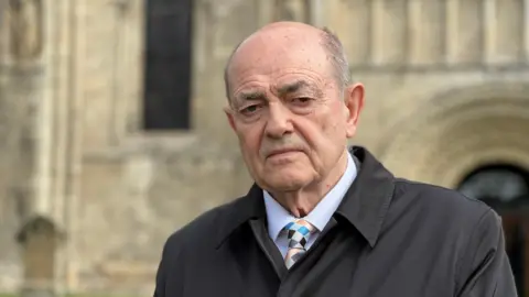 Michelle Lyons/BBC A bald man wearing a black coat, white shirt and colourful tie standing in front of a church