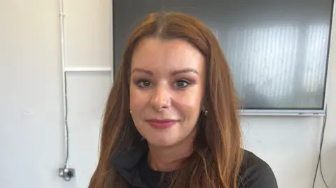 BBC A head and shoulders shot of Seana McConville who has long brown hair. She is wearing a dark top and has pink lipstick.