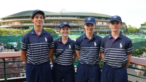 A group of four children wearing blue polo shirts with white hoops on the bottom. They have their arms behind their back and are smiling. They are stood in front of a stadium, with tennis courts also behind them.