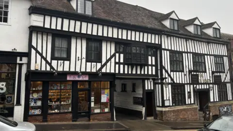 Katy Lewis/BBC The outside of a Tudor-style building, with white and black wooden timber frames and small windows. There is an alleyway in the middle, a hotel on the right hand side and a shop on the left.