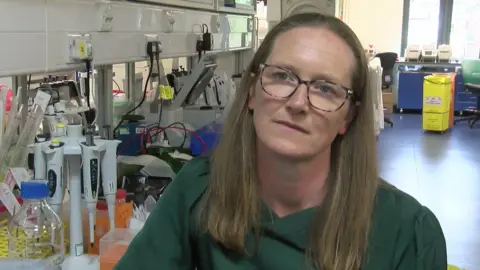 Professor Anne Corbett has long brown hair and black glasses. She is wearing a dark green shirt. She is sat in a hospital environment next to a desk full of equipment including microscopes and testing machinery. 