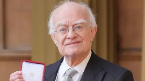 PA Media Sir John Rutter pictured at the Investiture ceremony holding his medal. He is wearing a suit with a black blazer, white shirt and silver tie. He has thin framed glasses, blue eyes and white hair. He is smiling at the camera.