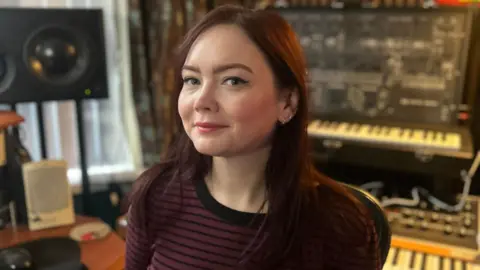 Handout Angie smiling looking at the camera. She has brown hair and is wearing a striped black and burgundy top. In the background, there are two keyboards and a speaker.