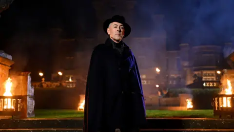 Peacock Alan Cumming standing in front of a castle at night, with various fires lit.  He is wearing either all black or purple, with a wide brimmed hat.  