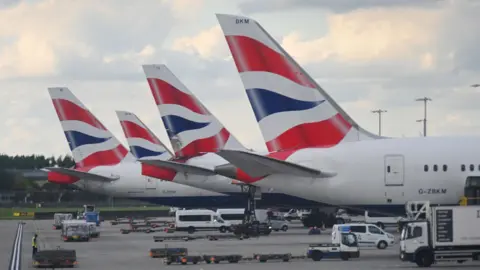 Four BA planes on the tarmac at Heathrow Airport. Only the tails of the planes are visible. They are white with red and blue livery on them.