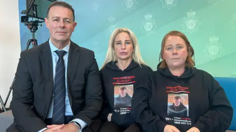 Ellis Cox's mother Carolyn is sat on a sofa with Det Ch Insp Steve McGrath to her right and and Julie O'Toole to her left. They are all looking at the camera. Carolyn has long blonde hair and Julie had mid length red hair. Both are wearing black hoodies with the writing "Who murdered Ellis Cox?" and a picture of Ellis. Det Ch Insp Steve McGrath is wearing a dark suit with a blue shirt and a navy blue tie with thin red stripes.