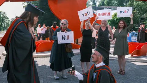 Steven Crichton Kat standing with her hands over her mouth looking at Steve on one knee holding a ring. Four people stand behind holding signs saying 'Will you marry me?'