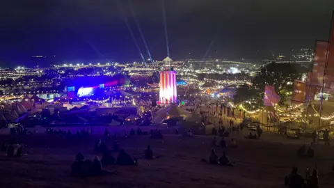 A view across Glastonbury Festival taken at night. There are lights as far as you can see. In the centre is the ribbon tower.