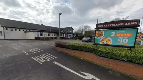 A sign with the words The Cleveland Arms is on the right hand side of the photo. A building goes from the left of the image to the middle, where a car is parked.