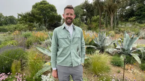 A man wearing a white shirt, mint green jacket and striped trousers stands amongst a garden featuring spiked plants and colourful flowers. He has short cropped brown hair, a beard and is wearing glasses.