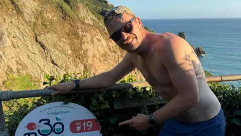 Matt Holland points at a sign for the 30 bays challenge, he is wearing a baseball hat, sunglasses and blue swim shorts. 