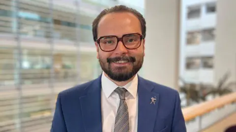 Deputy leader of the city council Ethan Radford wearing a blue suit with a blue and silver tie and glasses with a square brown frame.