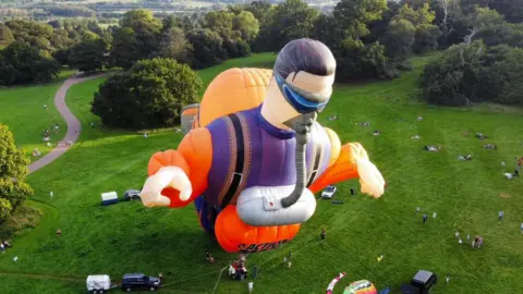 A giant hot air balloon shaped like Action Man tethered to the ground of a large field.