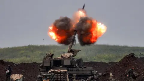 An Israeli self-propelled howitzer artillery gun fires rounds, with balls of fiery smoke around the weapon shooting into the air. The machine is surrounded by a dirt trench, with a grassy hill in front of it.