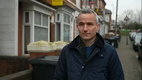 A BBC man with short gray hair wears a dark blue jacket and stands on a residential street with houses in the background.