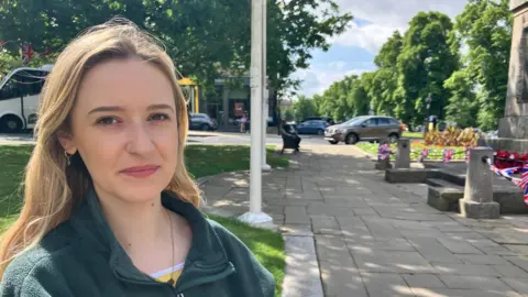 Caitlin Lee is sitting in the sunshine near Harrogate's war memorial. She is wearing a green fleece jacket and is looking straight at the camera.