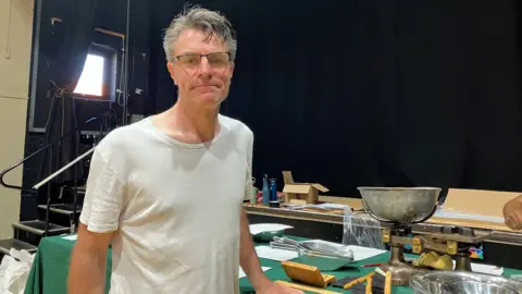James Yarker looks at the camera and stands in front of a table which has weighing scales on it and various measuring scoops. He is wearing a white t-shirt and has short grey hair.