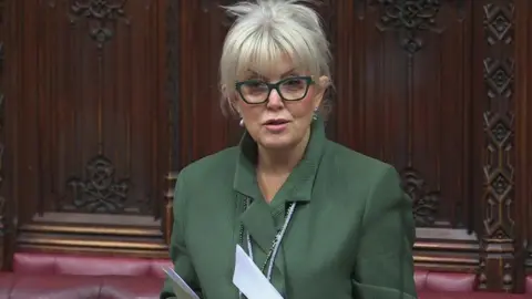 PA Media Baroness Newlove with blonde hair tied back, dark framed glasses and green jacket speaks during a House of Lords debate with wood panelling and a red leather bench in the background
