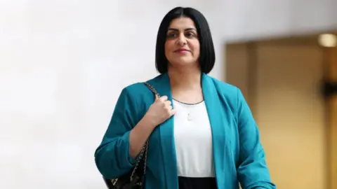 Mahmood is wearing a blue jacket and white top and has a short bob. She has a bag on her shoulder and is walking inside a building.