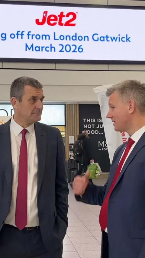 Two men in navy suits and red ties speaking in an airport.
