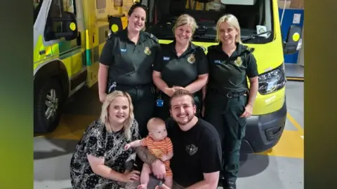 Jessica and Christian Roberts with baby Luca and three paramedics posing in front of an ambulance