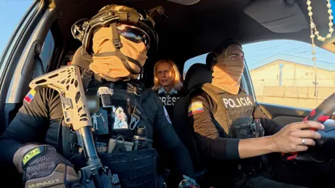 Two Ecuadorian police officers in dark sunglasses, helmets and balaclavas in a car with the BBC's Oral Guerin in the back seat