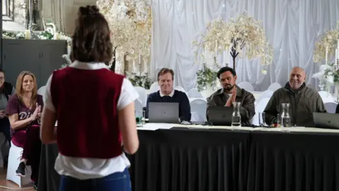 Three people are sitting behind a black table. They are looking towards a woman who is standing with their back towards the camera. She is wearing a red sweater vest and a white shirt.
