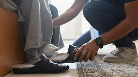 A nurse helps a person with their shoes at home. The close-up photograph shows a nurse crouching on the floor. The person receiving care is sitting on the bed wearing socks. Daylight is shining through the window and there is a patterned rug by the bed.