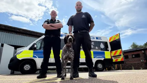 Kevin Shoesmith/BBC Two men, both wearing black police uniforms, stand with a brown and black dog in front of a police van.