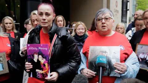 PA Media Two women stand in front of a larger group of people during the investigation into infected blood. They are both holding photos of their loved ones. The woman on the left has purple hair and is wearing a black down jacket over a red T-shirt. The woman on the right has short gray hair and wears glasses and a red T-shirt. A teal microphone with RTÉ written on it sits in front of her.