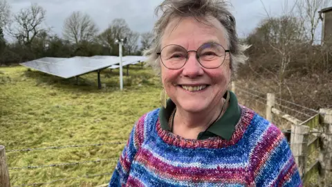 Lesley Bennett looks to camera, smiling. She is wearing a blue and purple striped jumper. Behind her is a field with solar panels in it.